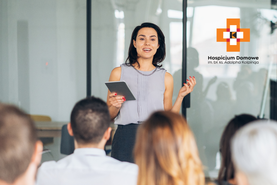 zdjecie przedstawiaj kobietę stojącą i przemawiającą do grupy osób siedzących na sali konferencyjnej.