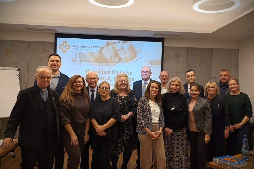 zdjecie grupowe uczestnikó zebrania - wszystkie osoy stoją uśmiechnięte na tle prezentacji z logiem Hospicjum